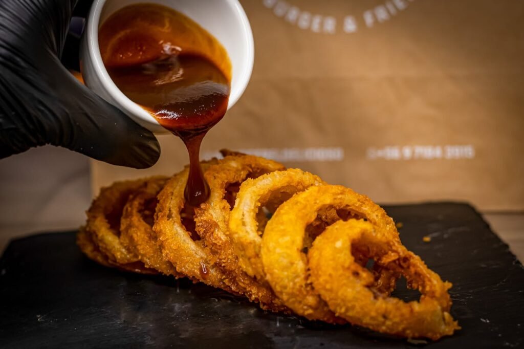 pexels-photo-17313960 Close-up of crispy onion rings with barbecue sauce being poured, highlighting delicious food photography.